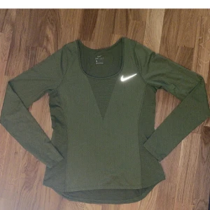 Grön långärmad träningströja Nike - Snygg grön långärmad träningströja från Nike med reflekterande logga på bröstet. Tillverkad i lätt och ventilerande Dri-Fit material som håller dig torr under träningspasset. Figurnära passform och rundad halsringning.