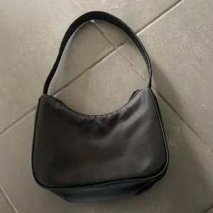 Svart axelväska i syntetmaterial - Enkel och stilren svart axelväska i syntetmaterial med mjuk form och smidig dragkedja upptill. Perfekt storlek för att bära det viktigaste och passar snyggt över axeln. Väskan har en minimalistisk look utan synliga loggor eller detaljer.