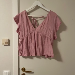 Rosa blus med knyt och volangärm - Supersöt ljusrosa blus från lager 157 med v-ringning, korta volangärmar och knyt i ryggen. Blusen är croppad och har en luftig, flowy passform i mjuk bomull. Perfekt för varma dagar när du vill ha något bekvämt men ändå stilrent. Katt finns i hemmet 