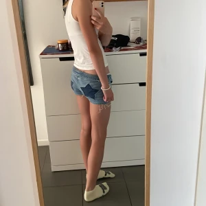Lågmidjade jeansshorts med stjärnor - Säljer ett par ljusblå lågmidjade jeansshorts med råa kanter och slitna detaljer. På baksidan finns två stora mörkblå stjärnor som ger en unik och cool look. Perfekta för sommaren och festivaler. Klassisk femficksmodell med knapp och dragkedja.