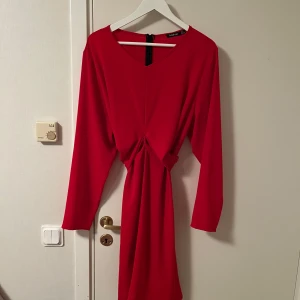 Röd långärmad klänning från Boohoo - Snygg röd klänning från Boohoo med lång ärm, v-ringning och knytning i midjan som ger en cool draperad effekt. Klänningen har dragkedja i ryggen och är i ett mjukt, lätt material som känns bekvämt hela dagen. Katt finns i hemmet 