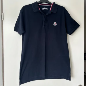 Mörkblå Moncler pikétröja - Snygg mörkblå pikétröja från Moncler med klassisk krage och knäppning framtill. Tröjan har diskret Moncler-logga på bröstet och rödvit detalj på kragen. Tillverkad i bomull. Passar M också.