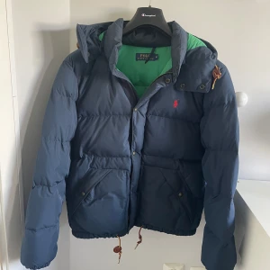  Polo Ralph Lauren jacka  - Snygg blå dunjacka från Polo Ralph Lauren med grön insida och röd broderad logga på bröstet. Jackan har huva, dragkedja och snörning i nederkant. Perfekt för kalla dagar och riktigt skön att bära.