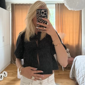 Svart croppad blus med knyt - Svart blus med korta ärmar och croppad passform. Toppen har en söt knytning framtill och lätt v-ringad hals. Perfekt att styla med höga jeans eller kjol för en trendig look.