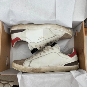 Pepe Jeans vita sneakers med röda detaljer - Snygga vita sneakers från Pepe Jeans med röda och bruna detaljer på hälen och tån. Skorna har snörning, rund tå och är tillverkade i skinn och textil. Perfekta för dig som gillar en clean men ändå sportig look. Det finns tecken på användning men ger ett bra hel hets intryck. Säljer då jag behöver få ihop lite pengar