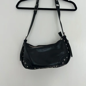 Svart axelväska med nitar - Cool svart axelväska med silvriga nitar längs kanterna och dragkedja upptill. Väskan har justerbar rem och extra dragkedjeficka på baksidan. Perfekt för dig som gillar snygga detaljer och vill ha en väska som sticker ut. Tyvärr är dragkedjan trasig, går förmodligen utmärkt att laga om man har koll!  