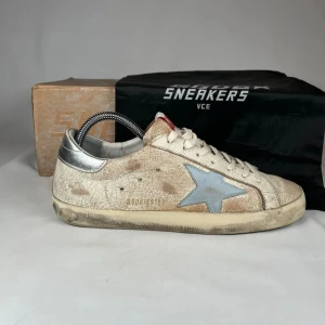 Golden Goose sneakers med blå stjärna - Snygga sneakers från Golden Goose med vintage-look. Skorna är beige med en ljusblå stjärna på sidan, silverfärgad häl och röda GGDB-detaljer på plösen. De har snörning och platt sula, tillverkade i skinn med slitna detaljer för en cool streetstyle.