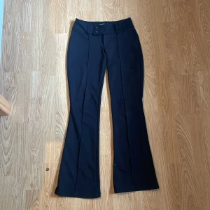 Svarta bootcut byxor från Gina Tricot - Snygga svarta bootcut byxor från Gina Tricot med pressveck och bred linning med dubbla knappar. Byxorna har en klassisk look och är tillverkade i ett mjukt material som ger en skön passform. Perfekta för dig som gillar stilrena och trendiga plagg.