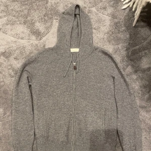 Grå kashmir hoodie från Sayless - Mysig grå hoodie från Sayless i lyxig kashmir. Tröjan har dragkedja framtill, huva och två fickor. Perfekt för dig som gillar en enkel men stilren look med premiumkänsla. Långärmad och supermjuk, passar till alla slags olika evenemang. OBS den är nopprig 