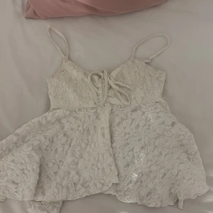 Vit spetsig linnetopp med snörning - Supersöt vit linnetopp med blommig spets och smala axelband. Toppen har snörning framtill som ger en snygg detalj och lätt utsvängd passform. Perfekt för sommarens varma dagar och ger en romantisk vibe.