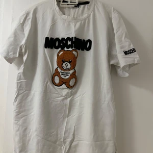 Vit Moschino t-shirt med nalle - Vit t-shirt från Moschino med broderad nalle och stor svart logga på bröstet. Även logotyp på ärmen. Klassisk rund hals och korta ärmar. Tillverkad i mjuk bomull, perfekt för en trendig och lekfull stil.