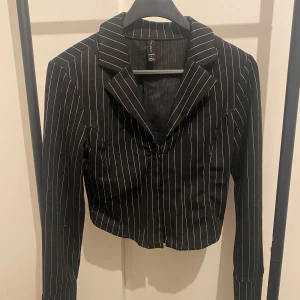 Svart randig kavaj från Amisu - Snygg svart skjorta från Amisu med vita tunna ränder. Modellen är croppad med klassisk krage och långa ärmar. Perfekt för dig som vill ha en stilren och trendig look. Materialet känns mjukt och följsamt.