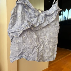 Ljusblå broderad linnetopp med volang - Supersöt ljusblå linnetopp med tunna axelband och volangdetalj framtill. Toppen har små vita broderade blommor över hela tyget och är perfekt för varma dagar. Luftig och lätt, passar till både jeans och kjol.