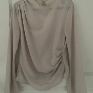 Beige blus - Säljer en ljusbeige blus med lång ärm som är vidare nertill, och snygg drapering på ena sidan så man får en form. Blusen har rund halsringning och en liten knapp i nacken. Tunt, flowigt material som ger en elegant look. Perfekt att styla med jeans eller kjol.