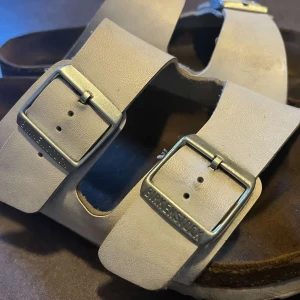 Beige sandaler från Birkenstock - Snygga beige sandaler från Birkenstock med två breda remmar i skinn och justerbara metallspännen. Klassisk korksula som formar sig efter foten och ger grym komfort. 