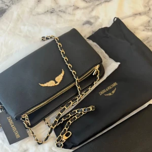 Svart axelväska från Zadig & Voltaire - Snygg svart axelväska från Zadig & Voltaire med guldfärgad kedja och dragkedja. Väskan har ett elegant guldigt emblem med vingar på framsidan och är tillverkad i skinn. Perfekt för dig som gillar stilrena detaljer och lyxig känsla.