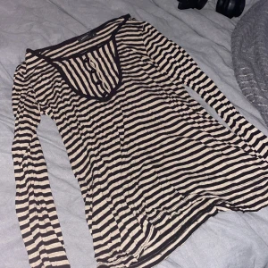 Randig långärmad tröja från Lindex - Säljer en svart och beige randig långärmad tröja från Lindex i storlek S. Tröjan har rund halsringning med knappar framtill och är i ett mjukt, tunt material som känns skönt mot huden. 