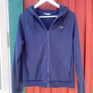 Blå zipup hoodie från Lacoste Sport - Mörkblå hoodie från Lacoste Sport med dragkedja hela vägen, huva med snörning och två fickor framtill. Klassisk krokodillogga på bröstet. Skön passform och ribbade muddar. 