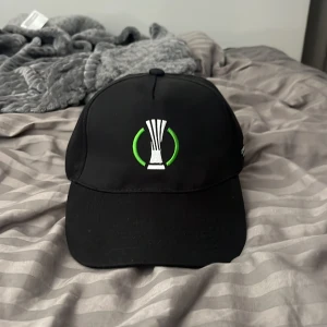 UEFA Conference League svart keps - Svart keps med broderad UEFA Conference League-logga framtill i vitt och grönt. Klassisk böjd skärm och justerbar passform. Perfekt för dig som älskar fotboll.