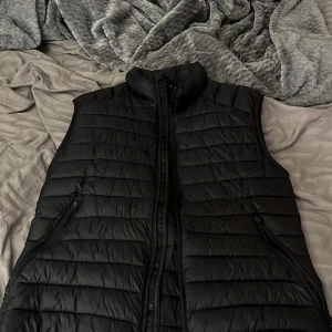 Svart dunväst från McKinley Small - Säljer en svart dunväst från McKinley i storlek Small. Västen har quiltad design, hög krage och dragkedja framtill samt två sidofickor med dragkedja. Yttertyg och foder är i nylon och fyllningen är polyester. 