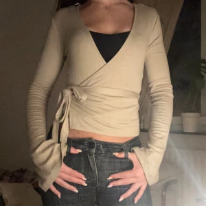 Beige omlottopp med knytband - Trendig beige omlottopp med långa ärmar och knytband i midjan. Toppen har v-ringning och är croppad, perfekt att styla med höga jeans. Ärmarna är lite utsvängda för en cool touch.
