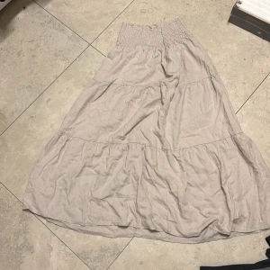 Beige långkjol från lager 157 - Luftig beige långkjol från lager 157 med smock i midjan och volangiga partier. Kjolen är tillverkad i ett mjukt bomullstyg och har en avslappnad passform som ger en skön och chill vibe. Perfekt för varma dagar när du vill ha något bekvämt och snyggt.