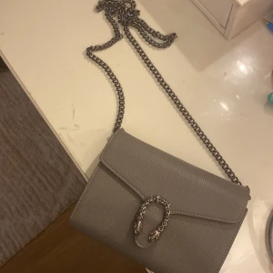 Grå axelväska med kedja och detaljspänne - Säljer en stilren grå axelväska i skinn med grov kedjerem och ett snyggt spänne framtill med detaljerade huvuden. Perfekt storlek för det viktigaste och ger en cool vibe till din outfit.