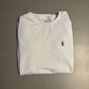 Vit t-shirt från Polo Ralph Lauren - Vit t-shirt från Polo Ralph Lauren med klassisk rund hals och broderad marinblå logga på bröstet. Modellen är slim fit och tillverkad i mjuk bomull. Perfekt basic-plagg med clean look och snygg passform.