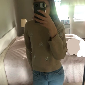 Beige stickad tröja med silverstjärnor - Beige stickad tröja med långa ärmar och coola silverglittriga stjärnor framtill. Baktill är den enfärgad beige! Tröjan har en avslappnad passform och ribbade muddar vid ärmslut och nederkant. Perfekt för dig som gillar en chill och trendig stil. 