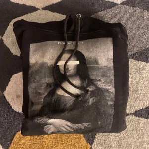 Svart hoodie med Mona Lisa-tryck - Svart hoodie med stor Mona Lisa-print framtill och snörning i huvan. Tröjan har långa ärmar, ribbade muddar och en liten vit detalj på ryggen. Perfekt för dig som gillar konstnärliga motiv. Passar dig runt 154-164 cm. Är från märket the cool elephant. Hör av dig vi några frågor💯