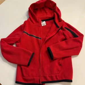 Röd Nike tech 122/128 - Nike hoodiejacka i rött med svarta detaljer och dragkedja. Jackan har huva, lång ärm och en svart rand över bröstet samt ficka med dragkedja på ärmen. Perfekt för dig som gillar sportig stil och vill sticka ut lite extra.