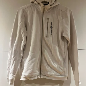 Vit hoodiejacka från Sail Racing - Vit hoodiejacka från Sail Racing med dragkedja framtill och bröstficka med logga. Jackan har huva med snörning och långa ärmar. Perfekt för dig som gillar en clean och sportig stil.