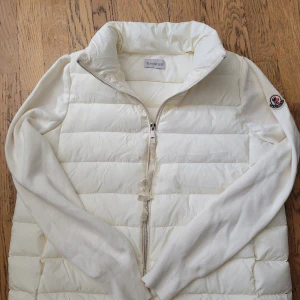 Vit dunväst med ärmar från Moncler - Snygg vit dunväst från Moncler med långa stickade ärmar och dragkedja framtill. Västen har hög krage och klassisk Moncler-logga på ärmen. Perfekt för lager-på-lager och stilren look. Storlek M men skulle också passa S.