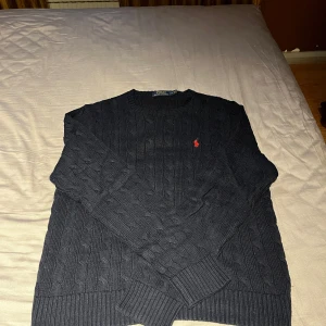 Mörkblå stickad tröja från Polo Ralph Lauren - Säljer en mörkblå stickad tröja från Polo Ralph Lauren med klassiskt kabelstickat mönster och röd broderad logga på bröstet. Tröjan har rund halsringning och långa ärmar. Perfekt för kyligare dagar! Storlek L men passar även M. Öppen för prisförslag!