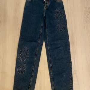 Mörkblå jeansbyxor med broderade mönster från Eytys - Unika mörkblå jeansbyxor från Eytys med vita broderade mönster på benen. Byxorna har raka ben, klassisk femficksdesign och knappar i gylfen. Perfekta för dig som vill sticka ut med coola detaljer.