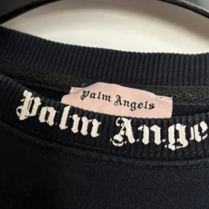 Svart sweatshirt från Palm Angels - Säljer en svart sweatshirt från Palm Angels med stor vit logotyp i gotisk stil över ryggen och nacken. Tröjan har rund halsringning och ribbade muddar. Perfekt för dig som gillar streetwear och vill sticka ut med en ikonisk design.