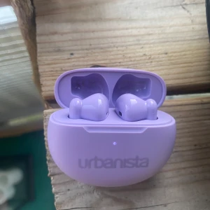 Lila trådlösa hörlurar från Urbanista - Säljer ett par snygga trådlösa hörlurar i lila från Urbanista. Kommer med matchande laddningsetui. Perfekt för musik, poddar och samtal på språng. Snygg och modern design med smidig förvaring i fodralet.