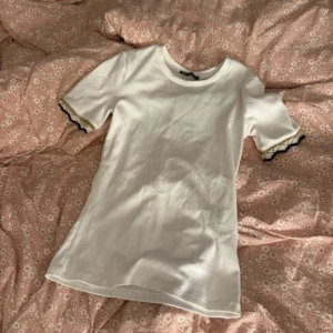 Vit topp med spetsdetalj från Zara - Säljer en vit topp från Zara med korta ärmar och svarta spetsdetaljer vid ärmsluten. T-shirten har en klassisk rund halsringning och är gjord i mjuk bomull. Perfekt för en stilren och söt look.💞💞