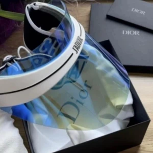 Dior J'Adior visir - Snyggt visir från Dior med blåtonat, transparent skärm och vit resår med svart text. Märkt med 'J'Adior' framtill och 'Christian Dior' på bandet. Perfekt accessoar för soliga dagar och trendiga outfits.