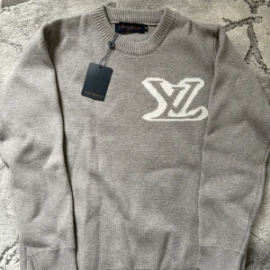 Grå stickad tröja från Louis Vuitton - Säljer en grå stickad tröja från Louis Vuitton med stort vitt LV-logo på bröstet. Tröjan har rund halsringning och långa ärmar. Perfekt för dig som gillar exklusiva märken och stilren design. Helt ny 