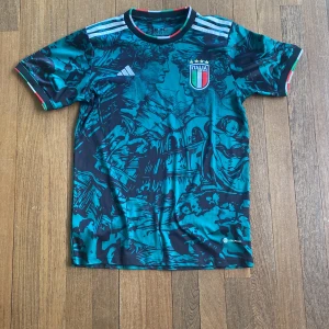 Italien fotbollströja Adidas - Säljer en blågrön Italien fotbollströja från Adidas med unikt konstnärligt mönster och broderat landslagsmärke på bröstet. Tröjan har korta ärmar, vita Adidas-ränder på axlarna och detaljer i Italiens färger vid ärmslut.