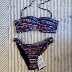 Färgglad bikini - Säljer en bikini med färgglatt mönster i blått, rött, vitt och lila. Överdelen har twistad front och knytband i nacken. Underdelen är klassisk med samma mönster. Perfekt för stranden eller poolen!