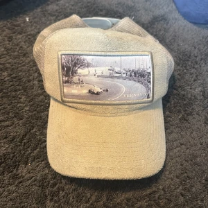 Beige keps från Vernando med racingmotiv - Snygg beige keps från Vernando med ett unikt svartvitt racingmotiv framtill. Kepsen har mesh på baksidan för extra ventilation och justerbar passform. Perfekt accessoar för en sportig och avslappnad stil.