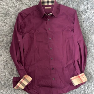Vinröd skjorta från Burberry Brit - Snygg vinröd skjorta från Burberry Brit med klassiskt rutigt mönster på insidan av kragen och manschetterna. Skjortan har långa ärmar, knappar framtill och är tillverkad i bomull med lite elastan för extra komfort.