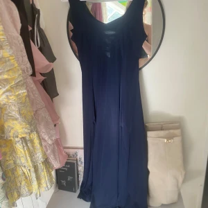 Marinblå långklänning med snörning i ryggen - Säljer en marinblå långklänning med snygg snörning i ryggen och v-ringad hals. Klänningen har en elegant slits framtill och är ärmlös, vilket ger en stilren och modern look. Perfekt för speciella tillfällen!