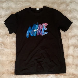 Svart t-shirt från Nike med färgglad logga - Svart t-shirt från Nike med en stor, färgglad logga framtill i blått, lila och rosa. Klassisk passform och rund halsringning. Perfekt för en sportig och avslappnad stil. Helt ny!