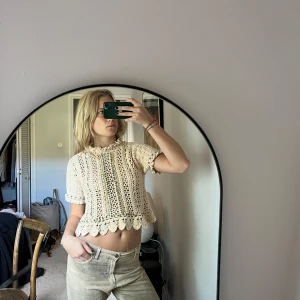 Beige virkad croppad topp från Zara - Superfin beige virkad croppad topp från Zara med korta ärmar och söt volangkant nertill och vid halsen. Toppen har ett diskret hålmönster och små gröna detaljer längs ärmslut och framtill. Perfekt för varma dagar!