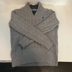 Grå stickad tröja från Polo Ralph Lauren - Säljer en klassisk grå stickad tröja från Polo Ralph Lauren med kabelstickat mönster och v-ringad hals. Har använts ett fåtal gånger och är i super bra skick. Pris kan diskuteras vid snabb affär!