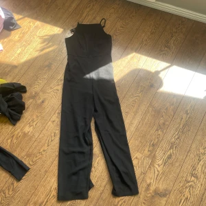 Svart byxdress med smala axelband - Snygg svart byxdress med smala axelband och hög halsringning. Vida ben och figurnära passform upptill ger en stilren och modern look. Perfekt för dig som vill ha något enkelt men ändå trendigt.