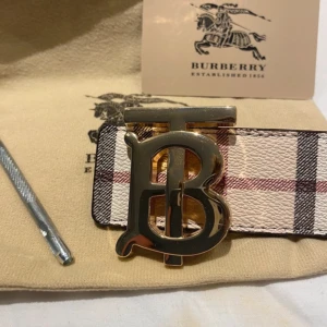 Burberry bälte med monogramspänne - Säljer ett Burberry bälte med klassiskt rutigt mönster i beige, svart och rött. Bältet har ett stort guldfärgat monogramspänne med bokstäverna TB. Perfekt accessoar för att lyfta din outfit.